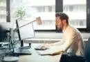 man in white dress shirt sitting on black rolling chair while facing black computer set and smiling