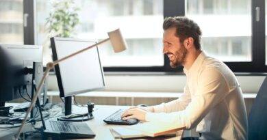 man in white dress shirt sitting on black rolling chair while facing black computer set and smiling